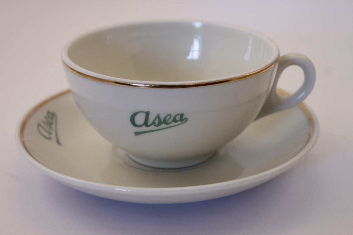 Kaffekopp med fat i cremevitt porslin med guld kanter och grön text: Asea på både kopp och fat.
