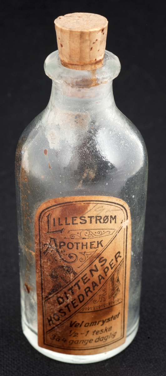 Rund glassflaske med korkpropp. Hvit dekorert etikett.