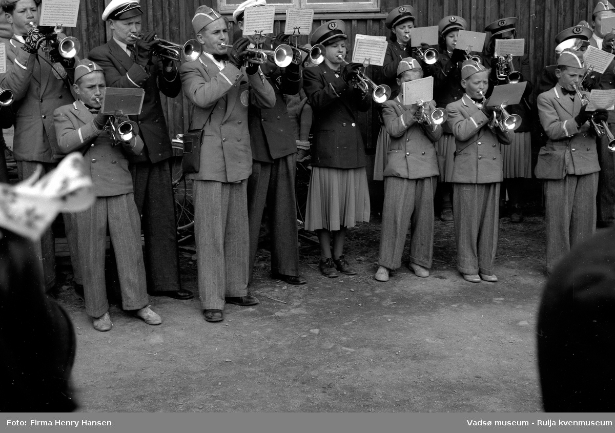 Vadsø sentrum 17. mai 1951. Musikkorps i uniformer, bestående av barn og voksne, står oppstilt og spiller. Vi ser en del av korpset, ikke dirigenten.  Korpset står foran postbrakka.