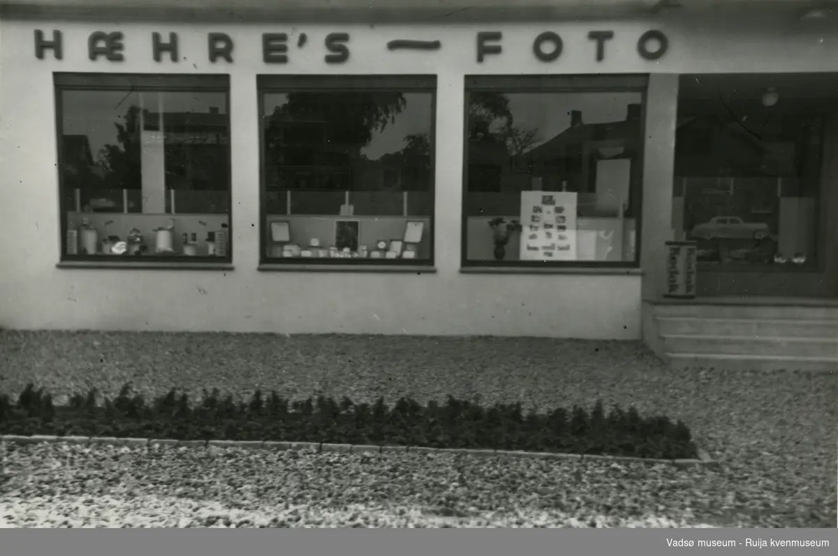 Forretningslokale. Fotobutikk med navn; "Hæhre's - foto". Første etasje med tre utstillingsvinduer og inngangsparti. I utstillingsvinduene ses fotoutstyr, fotorammer og en plakat med foto. På trappa inn til butikken står et reklameskilt for Kodak. Foran butikken er et blomsterbed.