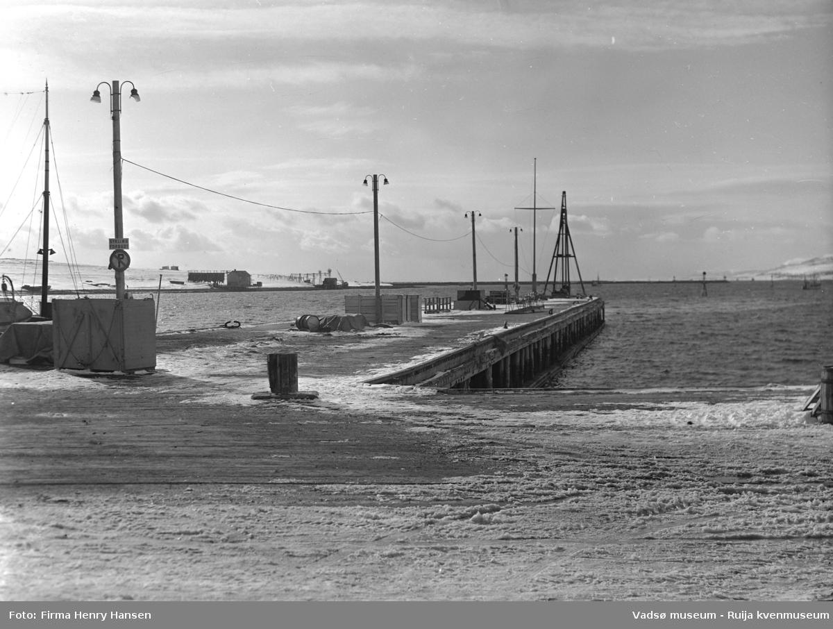Vadsø april 1952. Bildet er tatt fra dampskipskaien fra øst mot vest. Vi ser kaien og kaispissen med lysstolper, oljefat, et skilt med parkering forbudt/sykling forbudt og landgangen til hurtigruta. Til venstre i bildet ser vi Vadsøya og kaianlegg med kaihus, nothjul og hjellbruk bl.a. med fiskegarn til tørk. Lengst bort i bildet, mot vest, ser vi moloen som går fra Vadsøya mot nord og et par fiskebåter på havna.