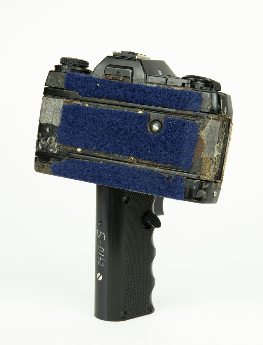 Handkamera HK 101 MT. På handkameran finns ett objektiv. Under kameran finns ett längre handtag varpå det finns 2 knappar för utlösning av film.