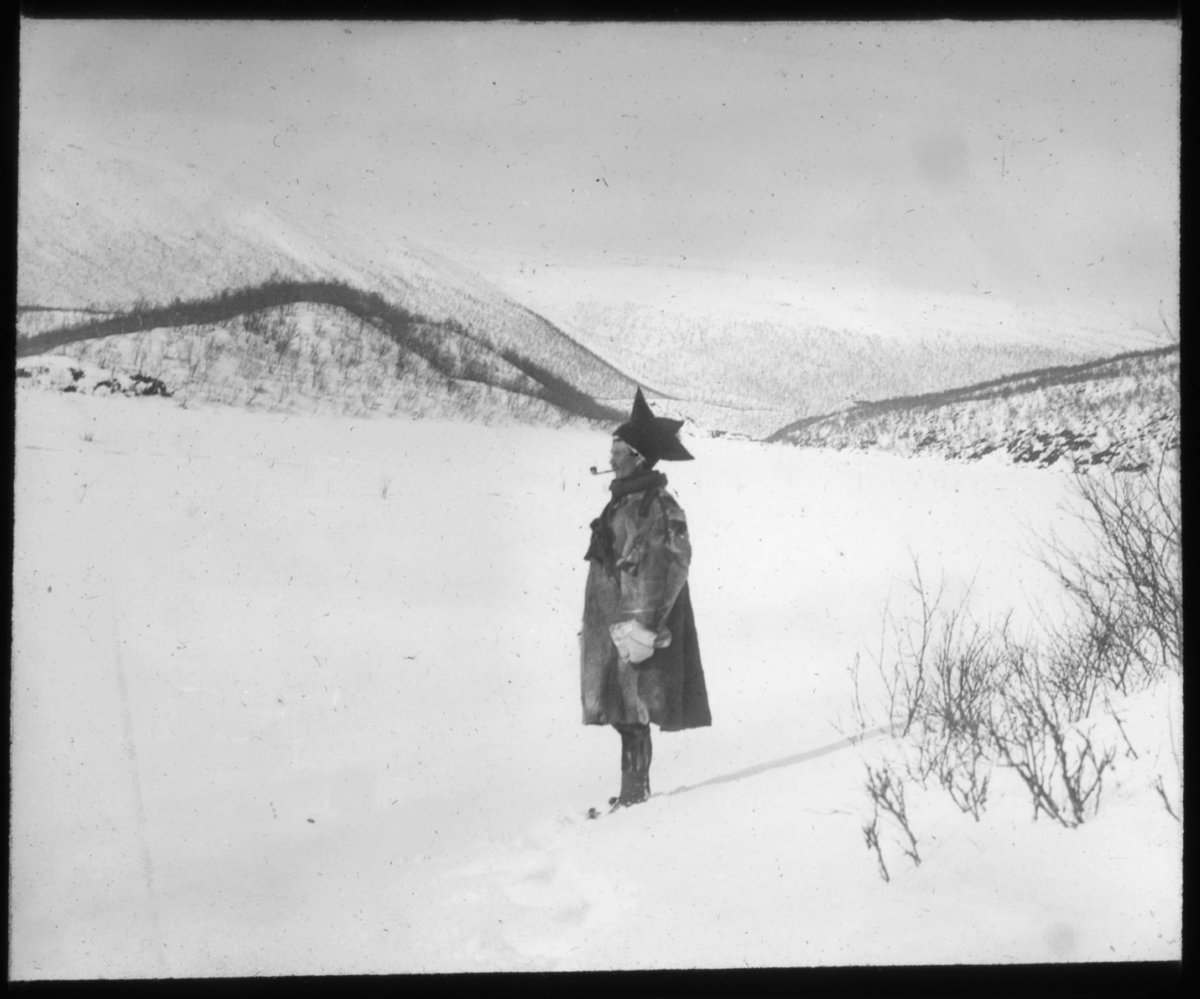 "Fjellsame. G.10" står det på glassplaten. Portrett av piperøykende same med pesk og samisk lue. Samen står oppreist i et vinterlandskap med mye snø og en fjellformasjon i bakgrunnen. Lua ser ut til å være fra Karasjok.