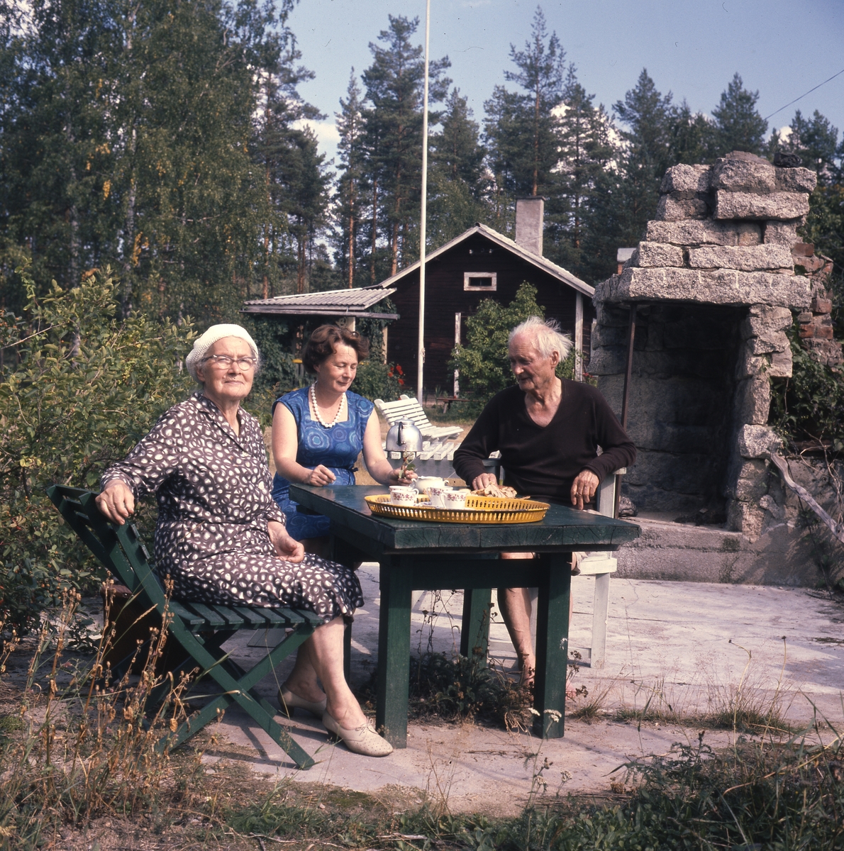 Vid Albert Vikstens skrivartorp. Några personer sitter i en trädgårdsmöbel och en dukad kaffebricka står på bordet. I mitten sitter Adéle Mickelsson och till höger Albert.
