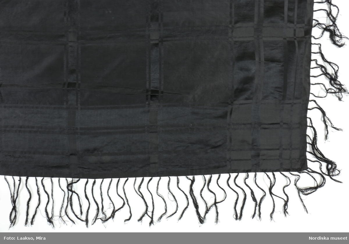 Kvadratiskt  stort halskläde av svart sidensatin  storrutig vävd i dräll, uppdelat på spegel och bård. Tråcklade fållar, iknuten frans av svart tvinnat silke.
I gott skick.
/Berit Eldvik 2011-11-28