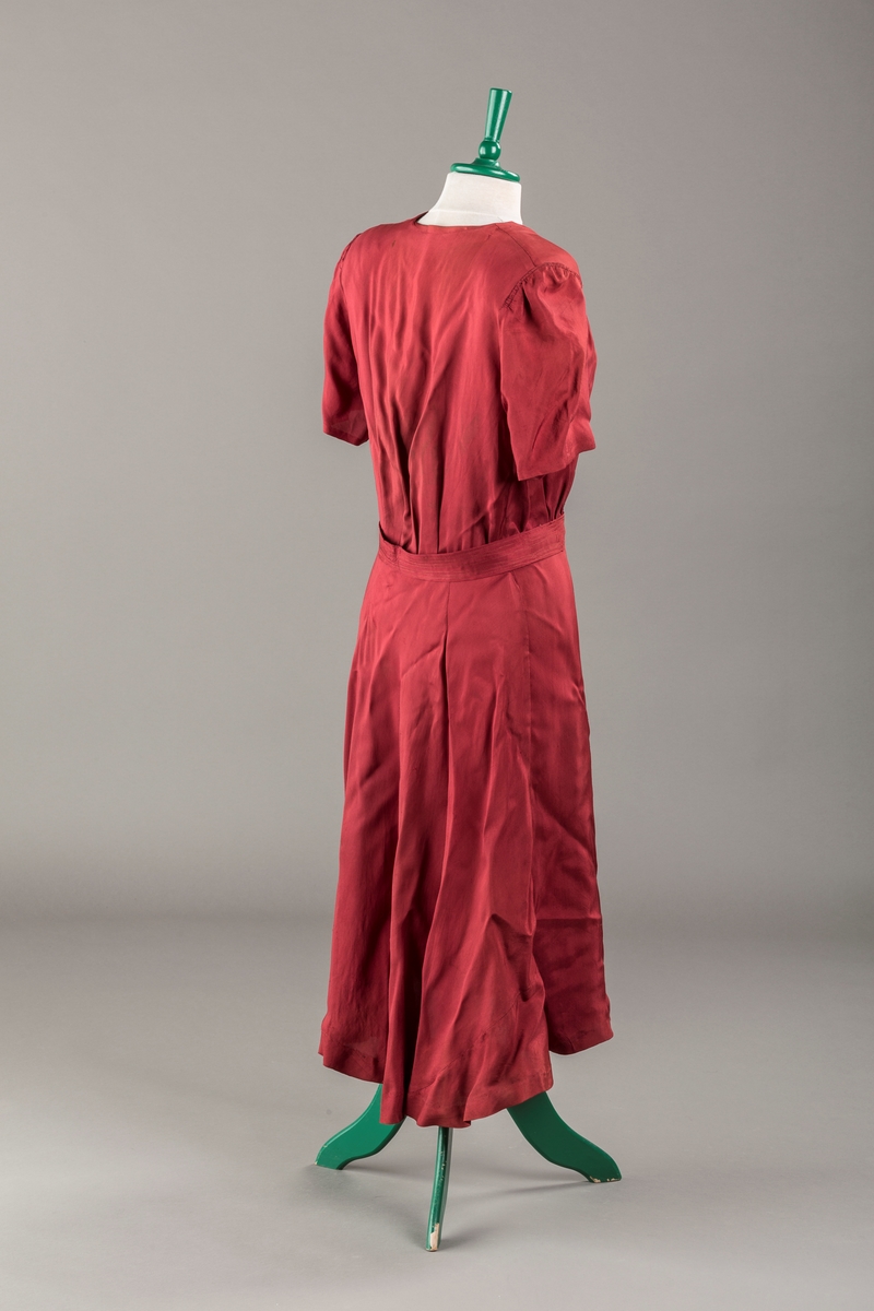 Form: Glatt, klokkeformet skjørt. Forstykkene rynket til buede bærestykker foran. Overdel rynket til skjørt. Korte ermer. Lukket med trykknapper. Belte fastsydd.