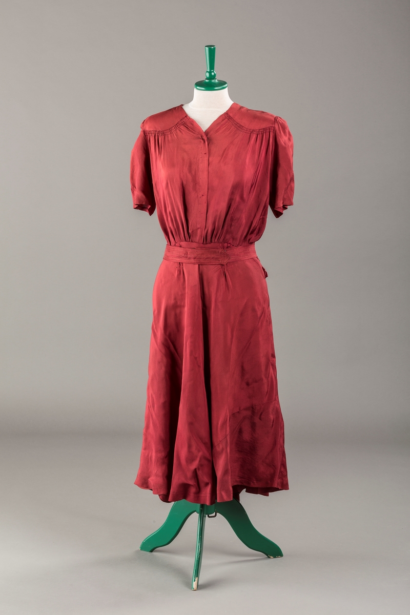 Form: Glatt, klokkeformet skjørt. Forstykkene rynket til buede bærestykker foran. Overdel rynket til skjørt. Korte ermer. Lukket med trykknapper. Belte fastsydd.