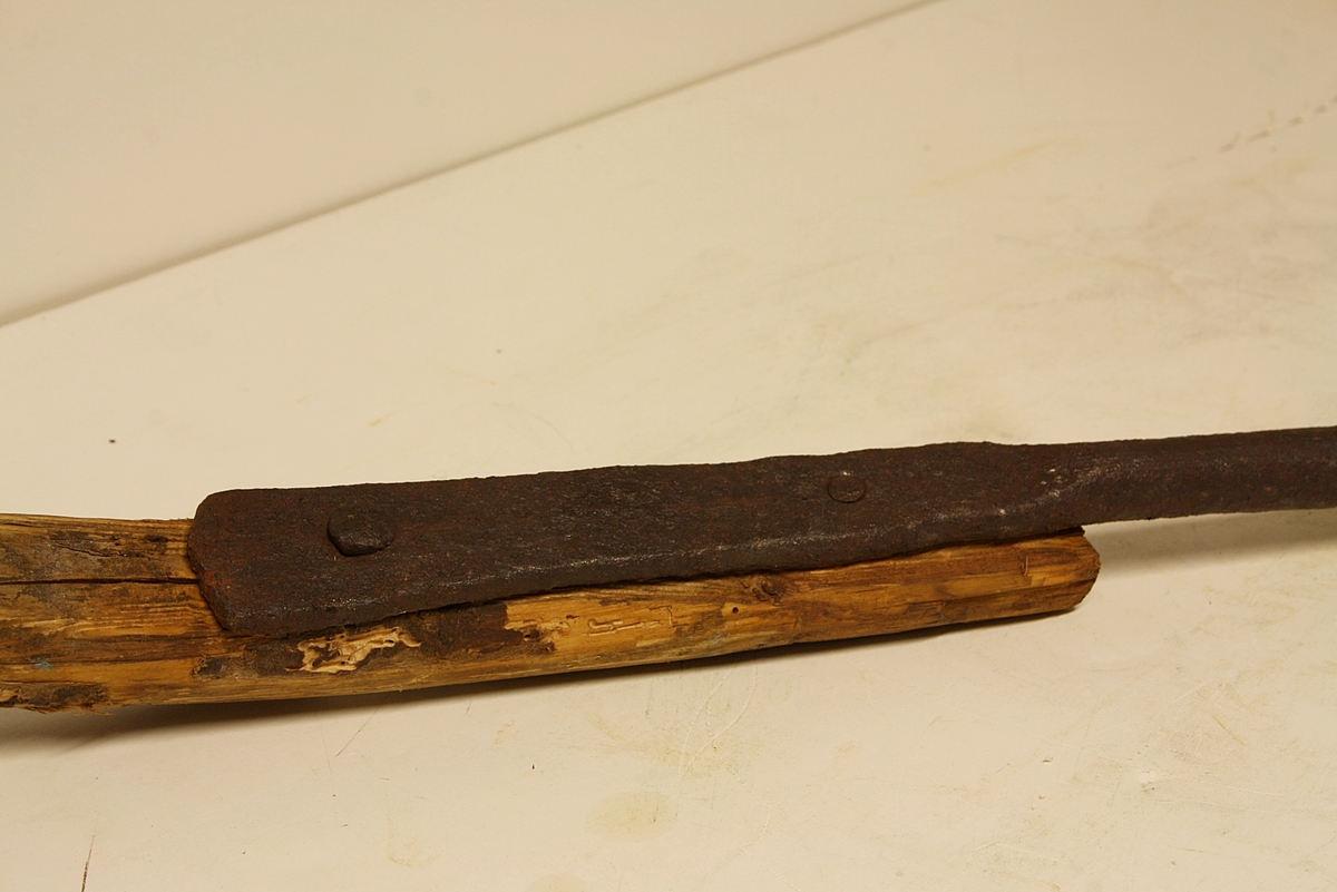 Grovt tilvirket treskaft. Langt metallskaft festet med to store spiker/nagler til treskaftet. Vifteformet, trekantet blad.