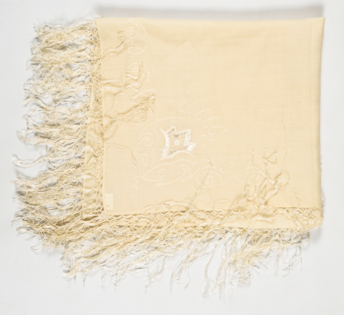 Tørkle i kremgul ullmusselin. Tamburert og brodert mønster med tynn lys silketråd i eine hjørnet. Påsette silkefrynser.
