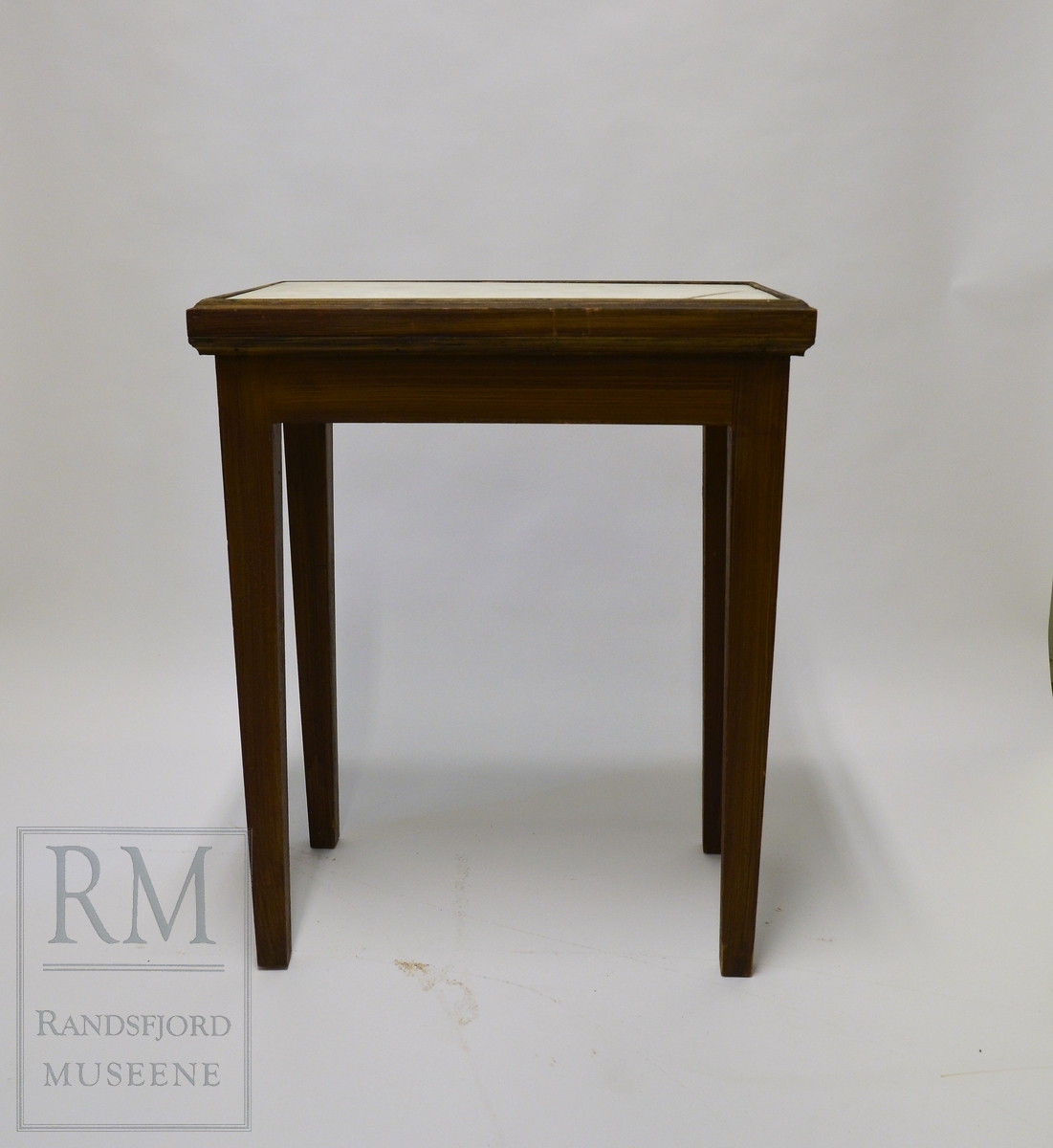 Lite bord, brunbeiset treverk. Bordplaten er opprinnelig en gravplate i marmor. Innskrift på undersiden. Et hjørne er sprukket.