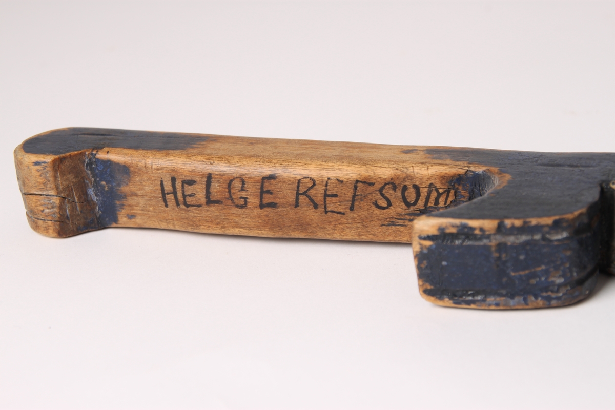 En firkantet skistav i tre. Øverste del av staven har et utskåret håndtak. Navnet til eieren Helge Refsum er skrevet på håndtaket. Nederst på tuppen av staven er det slått inn en liten metallstift.