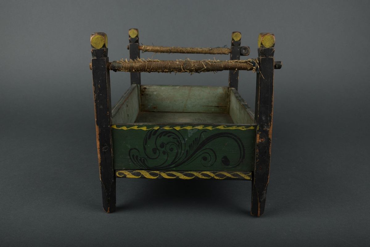 Dukkeseng i tre, med rosemalte sider og høye sengeposter.

De to øverste delene av sengen er surret med tau.