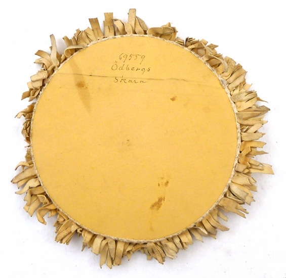 Lampmatta, rund mörkröd sammet i mitten, gråvita skinnremsor trädda på en virkad kant runtom, baksidan av gult bomullstyg, pappskiva inuti. 

Auktion 1933-05-13