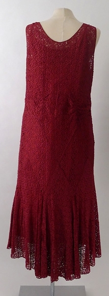 Lång klänning av röd silkesspets med underklänning av rött siden. Ett långt band i röd silkesspets är fastsytt vid vardera axel framtill på klänningen.