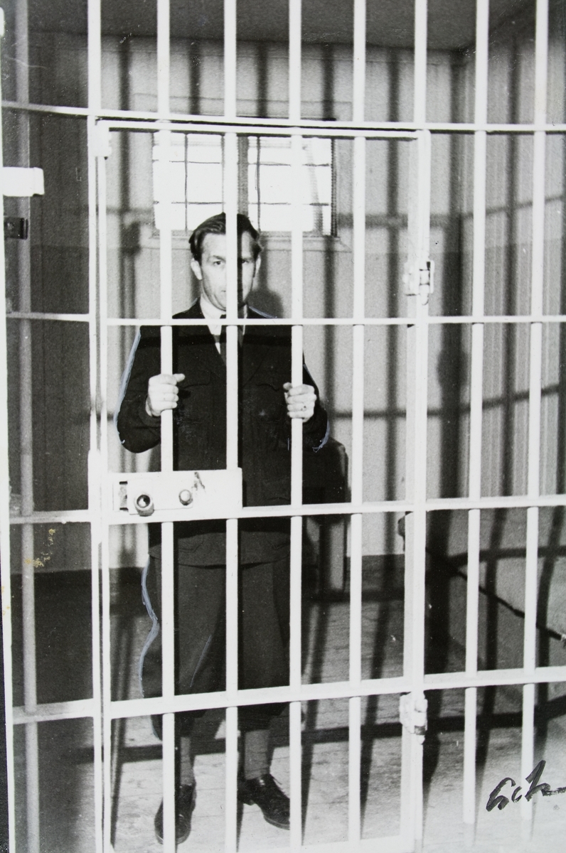En mann står inne i en fengselscelle, bak et gitter. Bildet er tatt i Trondheim kretsfengsel, avd. B i i Munkegata 16.