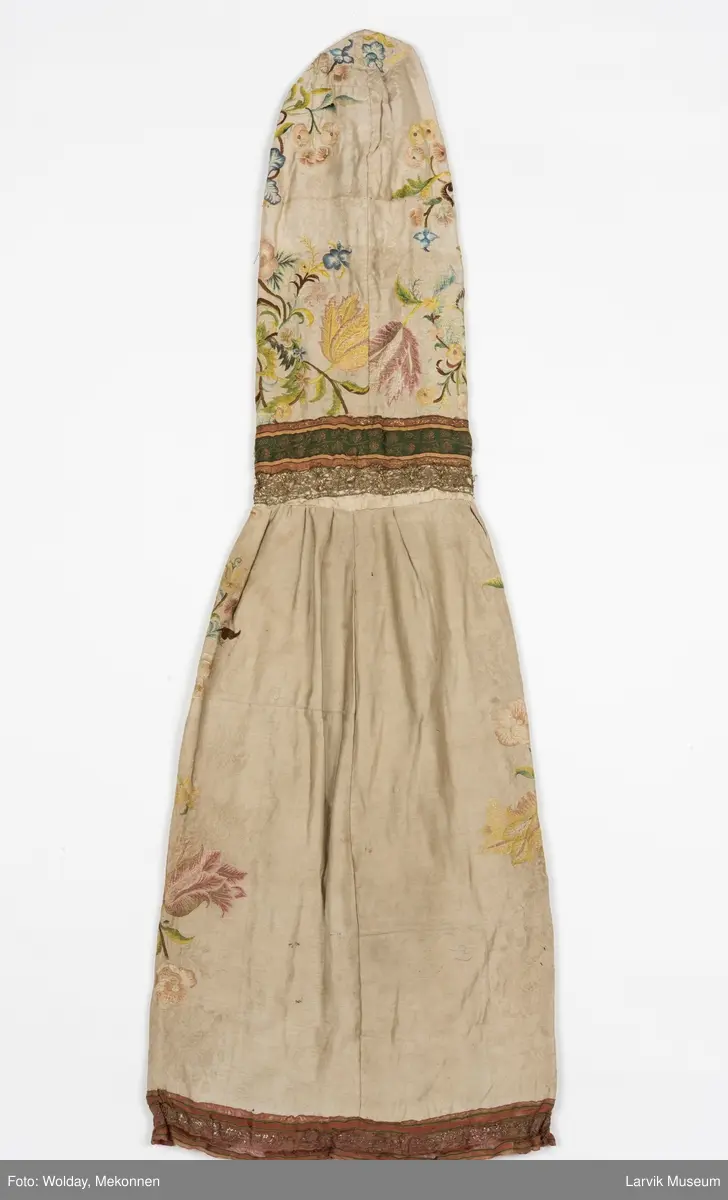 Form: livstykke som hette, åpen til livet, bred linning, rynket skjørt med blondekant nederst, søm bak på hetten i linning, søm foran og bak i skjørt, enkelte skjøtesømmer, linning brodert med gulltråd, skjørtet er foret med tynt stoff