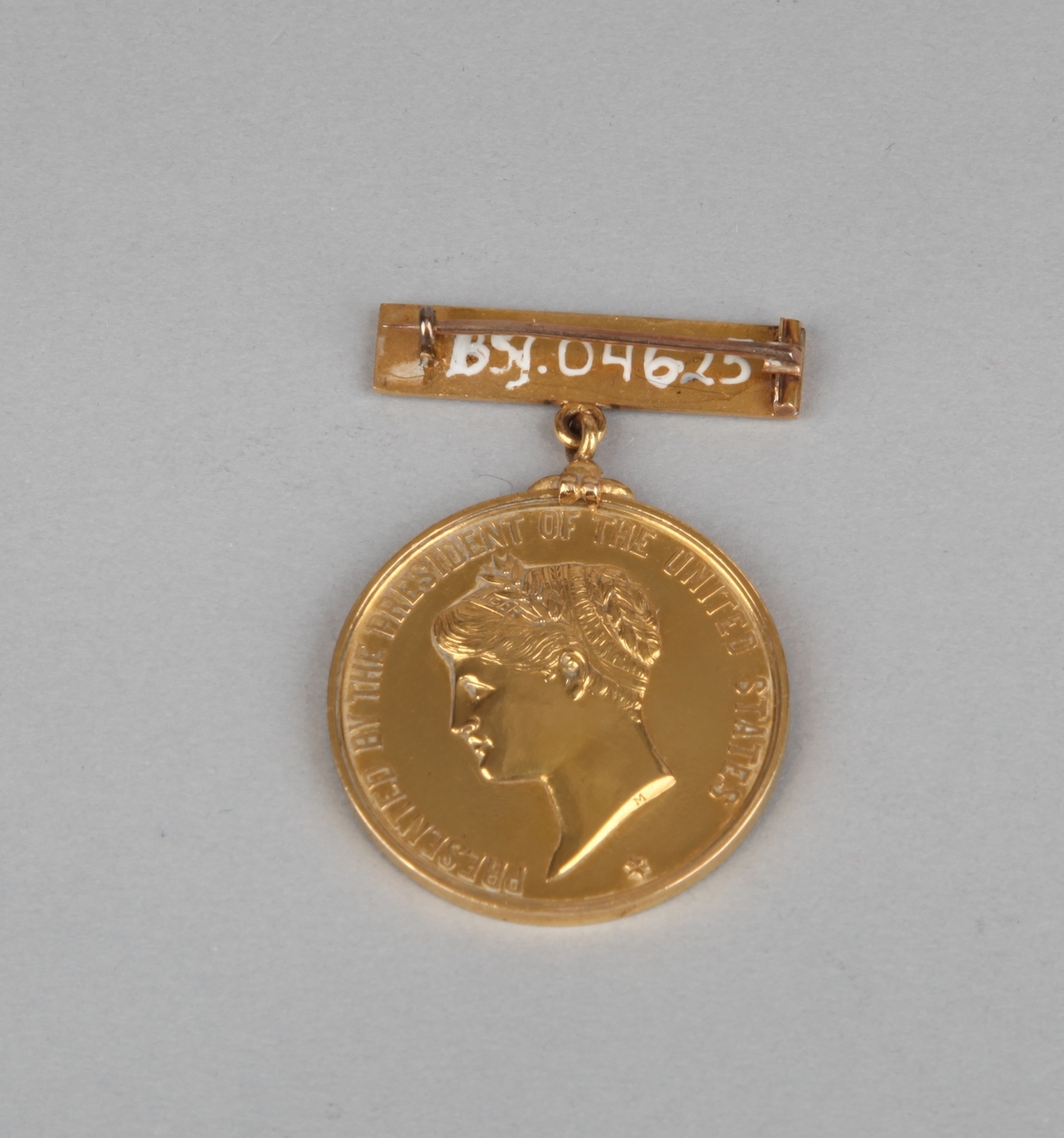 Rund gullmedalje med nål med motiv av kvinnehodeog tekst på forsiden samt tekst på nålfeste og baksiden.