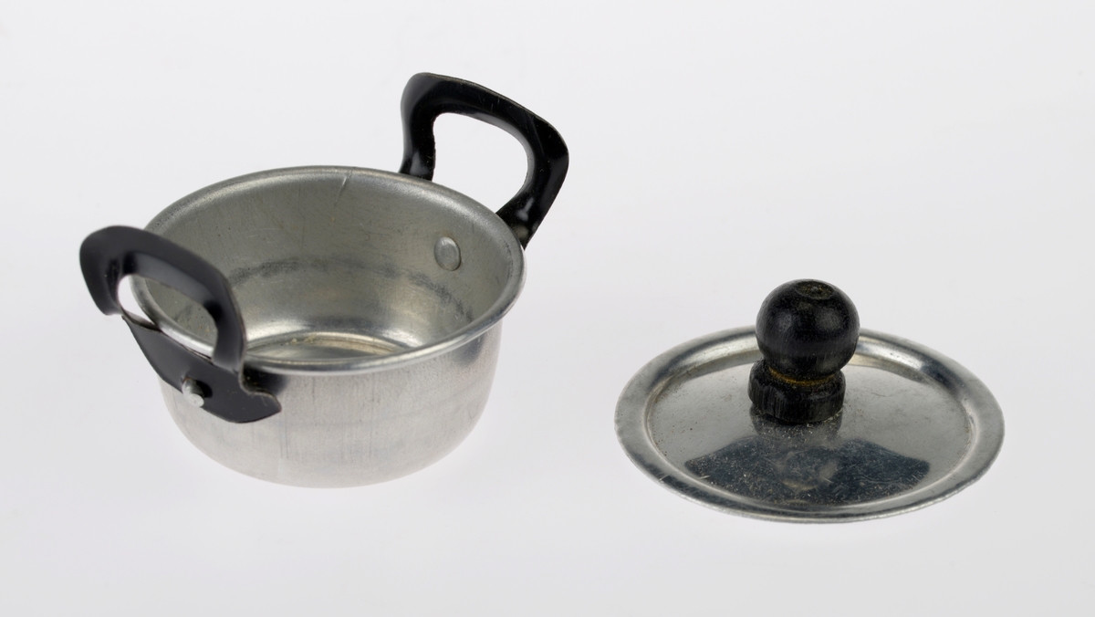 Et kjøkkensett som er montert i en liten pappeske med lokk. Settet er trolig fabrikkprodusert. Gjenstandene er undernummerert 01-10.
FTT.54457.01: En kasserolle med lokk laget av blikk. I hver side av kasserollen er det relativt store svartmalte håndtak av metall, og på lokket er det en treknott som også er svartmalt. Håndtakene og knotten er loddet på. Kasserollen er trolig valset.
FTT.54457.02-03: To identiske tallerkener laget av blikk. De er runde med en bred rand.
FTT.54457.04-05: To identiske kniver laget av metall, og de er antageligvis støpt. Håndtaket har prikker langs kanten som står opp fra metallet, og i midten er det en opphøyet stripe av metall. Knivbladet er relativt bredt og avrundet i tuppen. 
FTT.54457.06-07: To identiske skjeer laget av metall, og de er antageligvis støpt. Håndtaket har prikker langs kanten som står opp fra metallet, og i midten er det en opphøyet stripe av metall. 
FTT.54457.08-09: To identiske gafler laget av metall, og de er antageligvis støpt. Håndtaket har prikker langs kanten som står opp fra metallet, og i midten er det en opphøyet stripe av metall. Gaflene fire tenner.
FTT.54457.10: Esken der gjenstandene 01-09 er montert i. Esken og lokket er laget av papp og er stiftet i hjørnene. Esken er grå og blå på farge. Langs kanten inni esken er det klippet ute en tungekant. I bunnen er det en rød papplate der det er klippet ut hull for montering av de ulike gjenstandene.