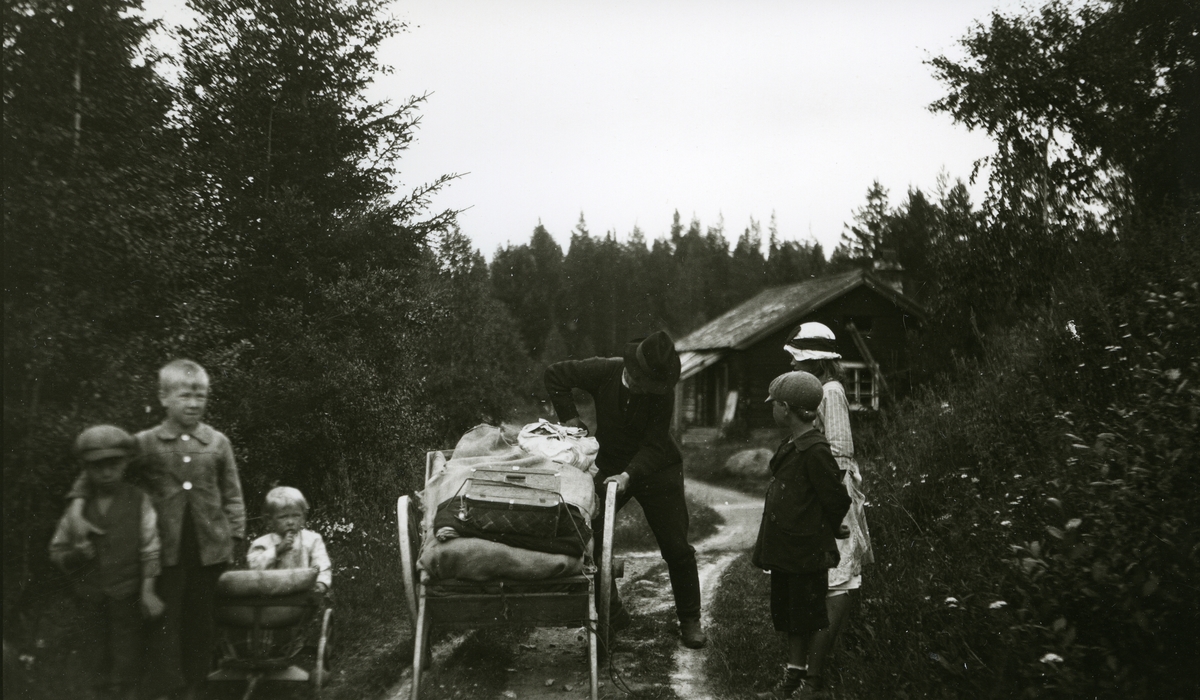 Mann m/kjerre ved Horrmundbruket, Dalarne (Sverige). Fem unger står rundt ham. Lita "stue" (hus) i bakgrunnen