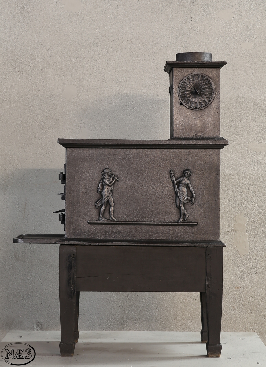 Kokeovn med to muser på enkel profillist. En fløytespillende og en dansende. Begge med draperier. Kokeanretning i topplaten til 1.etg. Ovnsdør på kortside. Rosett på begge sideplatene 2.etg. Den ene er løs og fungerer som ventil. Innskrift på frontplate og bakplate 2.etg. Røykuttak i toppen.