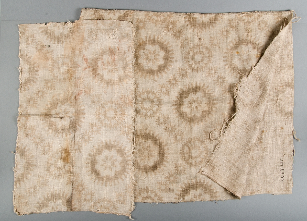 Två fragment från matta. Grov tuskaftad linneväv med tryckt eller schablonmålat brunt mönster. Den större biten hopsydd av två våder med stadkanter, en sida fållad.
