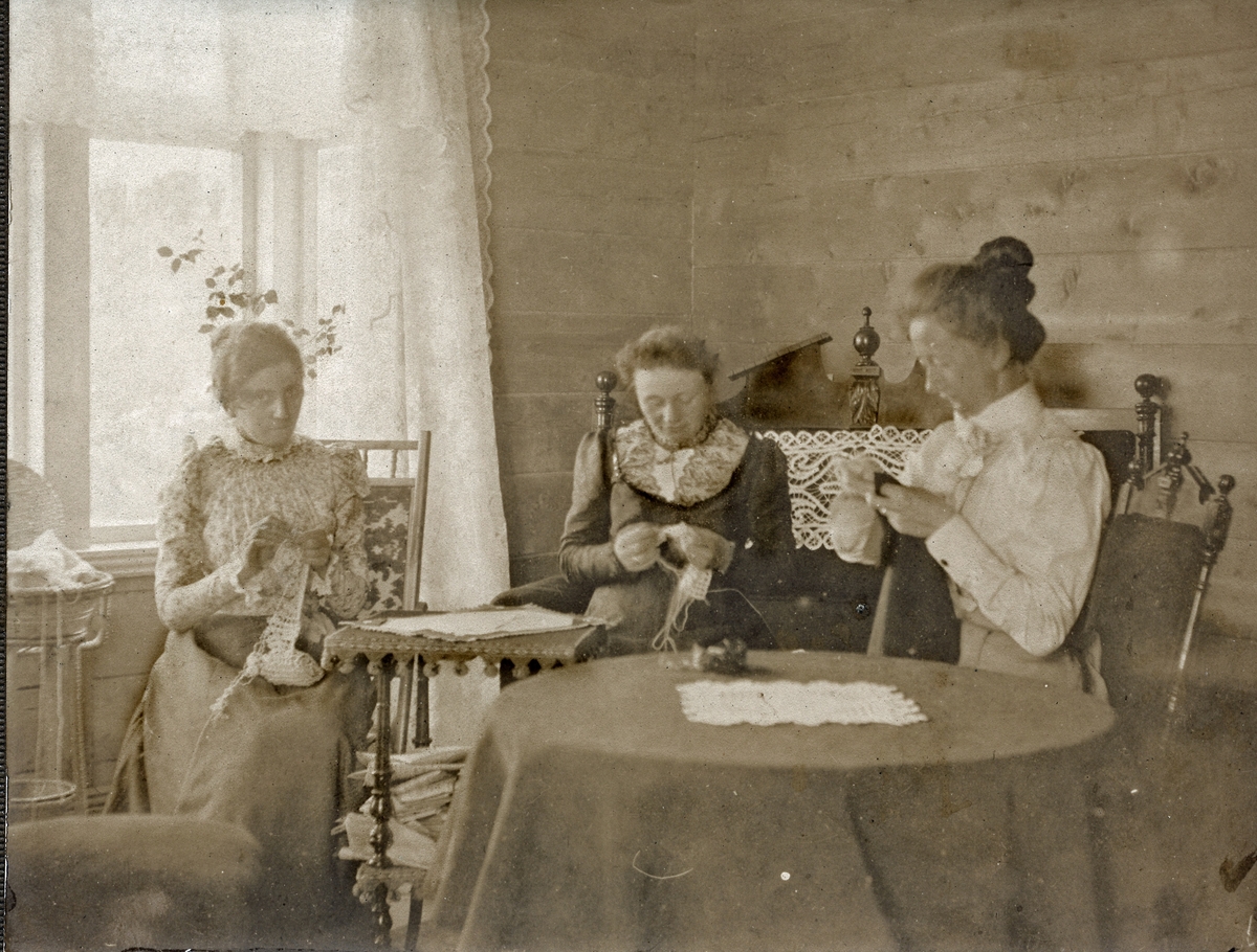 Gruppe kvinner. Tre kvinner sitter med håndarbeid (broderi eller hekling) ved vindu. Nanna Pedersen, fru Laws, frk. Aabakken. Interiør. Umalte plankevegger.