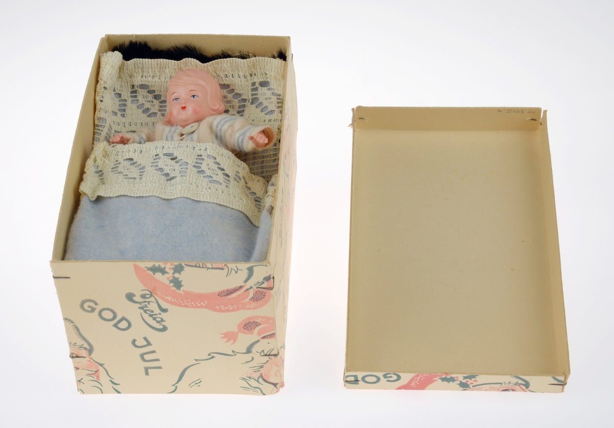 En sjokoladeeske brukt som dukkeseng med dukke, dyne, pute, skinnfell, ullteppe og madrass. Gjenstandene er undernummerert 01-08.
FTT.54432.01: En sjokoladeeske fra Freia brukt som dukkeseng. Esken er med lokk og laget av papp. Den er stiftet i hjørnene. Utenpå er det trykt julenissehoder i rødt og grønt. For tekst se "Påført tekst/merker".
FTT.54432.02: En dukke laget av plast der både armer og bein er bevegelige ved at de er festet med strikk inni dukken. Håret er støpt og er krøllet ved ørene. I ansiktet er det malt øyebryn, øyne, røde kinn og munn. Dukken har på seg en pyjamas laget av flanell. Den er stripete i fargene hvit, blå, lys oransje og grønn. På skjorten er det to knapper i fronten.
FTT.54432.03: En hodepute laget av blått bomullsstoff med heklet trekk. Puten er fyllt med diverse tekstilbiter.
FTT.54432.04: En dyne laget av blått bomullsstoff med en påsydd blondekant foran. Baksiden er sydd sammen av tre ulike tekstiler. Dynen er fyllt med tekstilbiter.
FTT.54432.05: En brunsvart skinnfell, trolig laget av kaninpels.
FTT.54432.06: Et ullteppe som trolig klippet ut av et eksisterende ullteppe. Teppet er gulhvitt med blomster og blader i rosalilla.
FTT.54432.07: Et sammenbrettet stykke som er heklet. Trolig en rest som er brukt som madrass i esken/sengen.
FTT.54432.08: Et sammenbrettet stykke som er heklet. Trolig en rest som er brukt som madrass i esken/sengen.