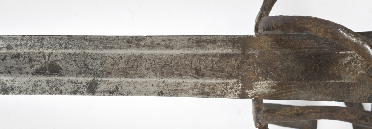 Avleng, plommeformet knapp med nitteknapp. Tregrep med lær bevikling. Asymmetrisk feste med flate bøyler. Fremre parerstang går over i håndbøylen. Sidering. 
Tvenegget, rett klinge med elipseformet tverrsnitt. og 1 kort midtstillt  blodrand på hver side.. Klingen er merket med  Passau-ulv på utsiden og smedmerke på innsiden.