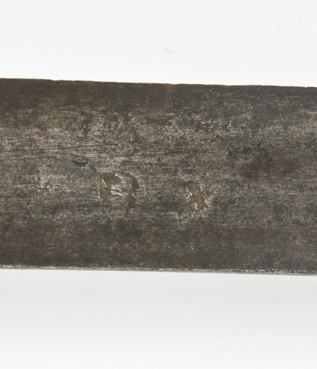 Pyramideformet knapp med strekdekor. Tregrep med hyssing bevikling. Runde parerstenger med filt strekdekor og svakt opp- og nedbøyde ender. Kurvfeste med runde bøyler med utsmidde, skråfilte midtpartier. Nyreformet parerplate med tombøyle på innsiden 2 sidebøyler på innsiden..
Enegget, krum klinge med bred blodrand. Gjeddenebb og tveegget mot spissen
Klingens innside merket med tre smedstempeler hvorav et er MR