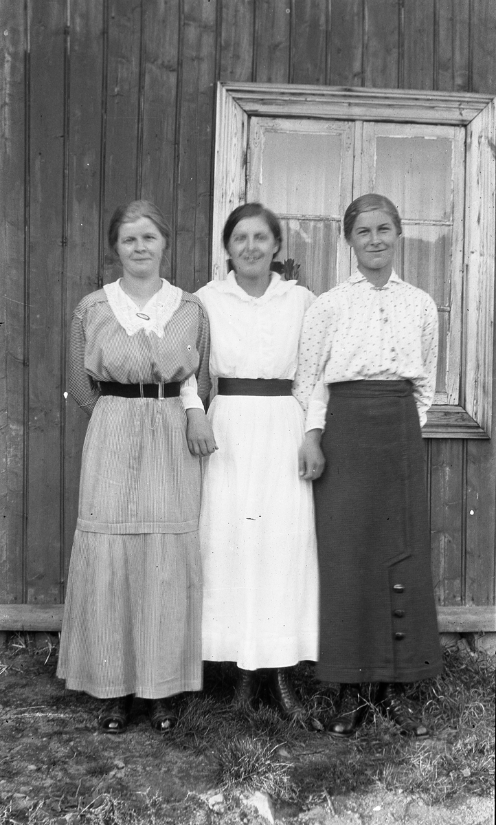 Tre kvinner i helfigur ved en husvegg. I midten Signe Hagen som konfirmant, til høyre trolig søstera Nora Hagen, og til venstre trolig Olga Hagen, også søster. Stedet er sannsynligvis Nordeng i Øverskreien.