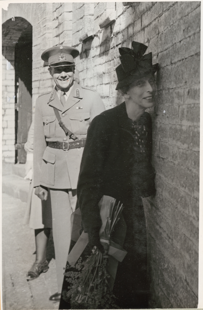 Drottning Louise, klädd i mörk dräkt och hatt, står framåtlutad mot murverket tillhörande Kungsholms fort. I handen har hon en blombukett. Bakom drottningen står en uniformsklädd militär från armén och ler.