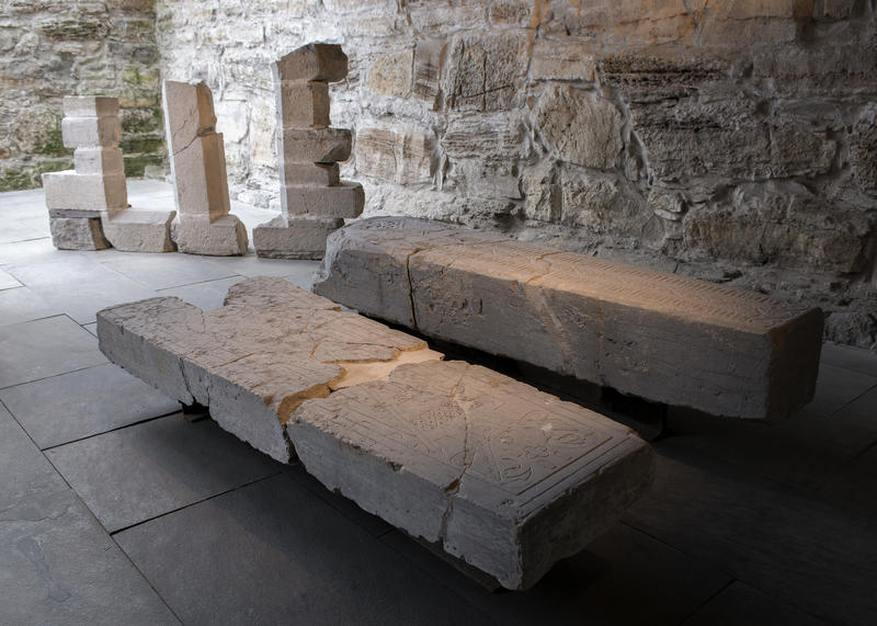 To middelaldergravsteiner ligger utstilt i Domkirkeoddens lapidarium, med steinramma til et gotisk vindu i bakgrunnen.