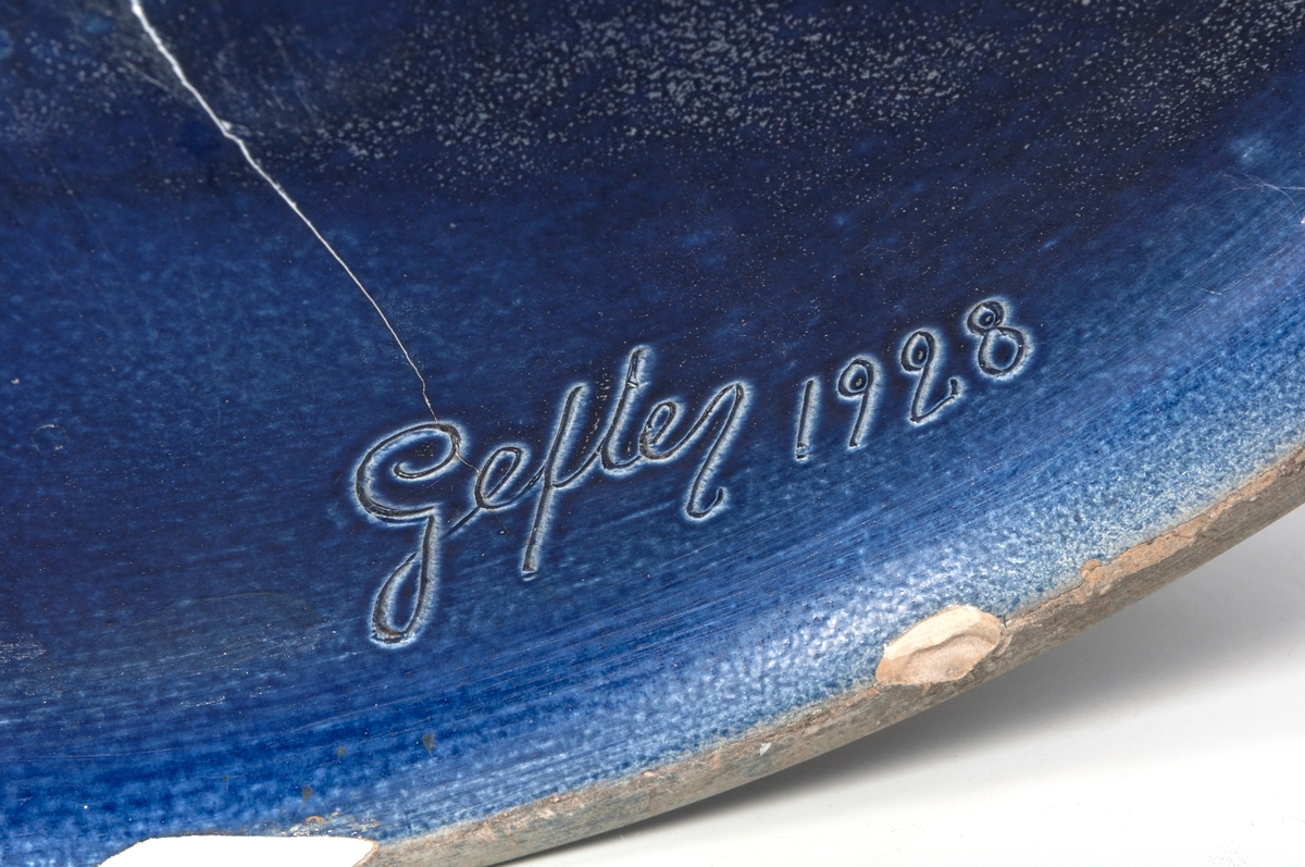 Trädgårdsurna i blåglaserad keramik, tillverkad 1928 av Gefle Porslinsfabrik, formgivare Arthur Percy. Klassisistisk formgivning. Skulpturerad reliefdekor, en på varje sida, föreställande en kvinna på delfin. Märkt i botten "Gefle 1928".