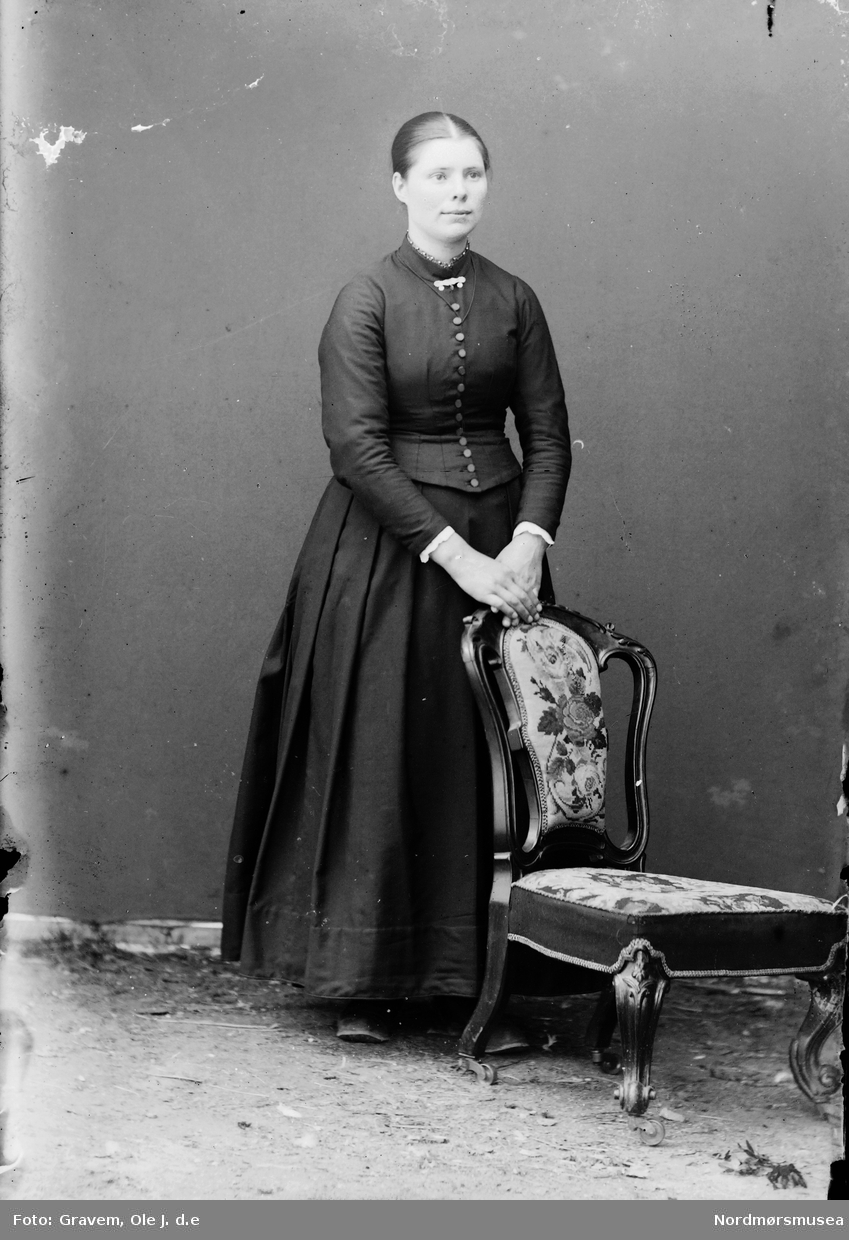 Portrett i helfigur av ung kvinne stående ved stol.