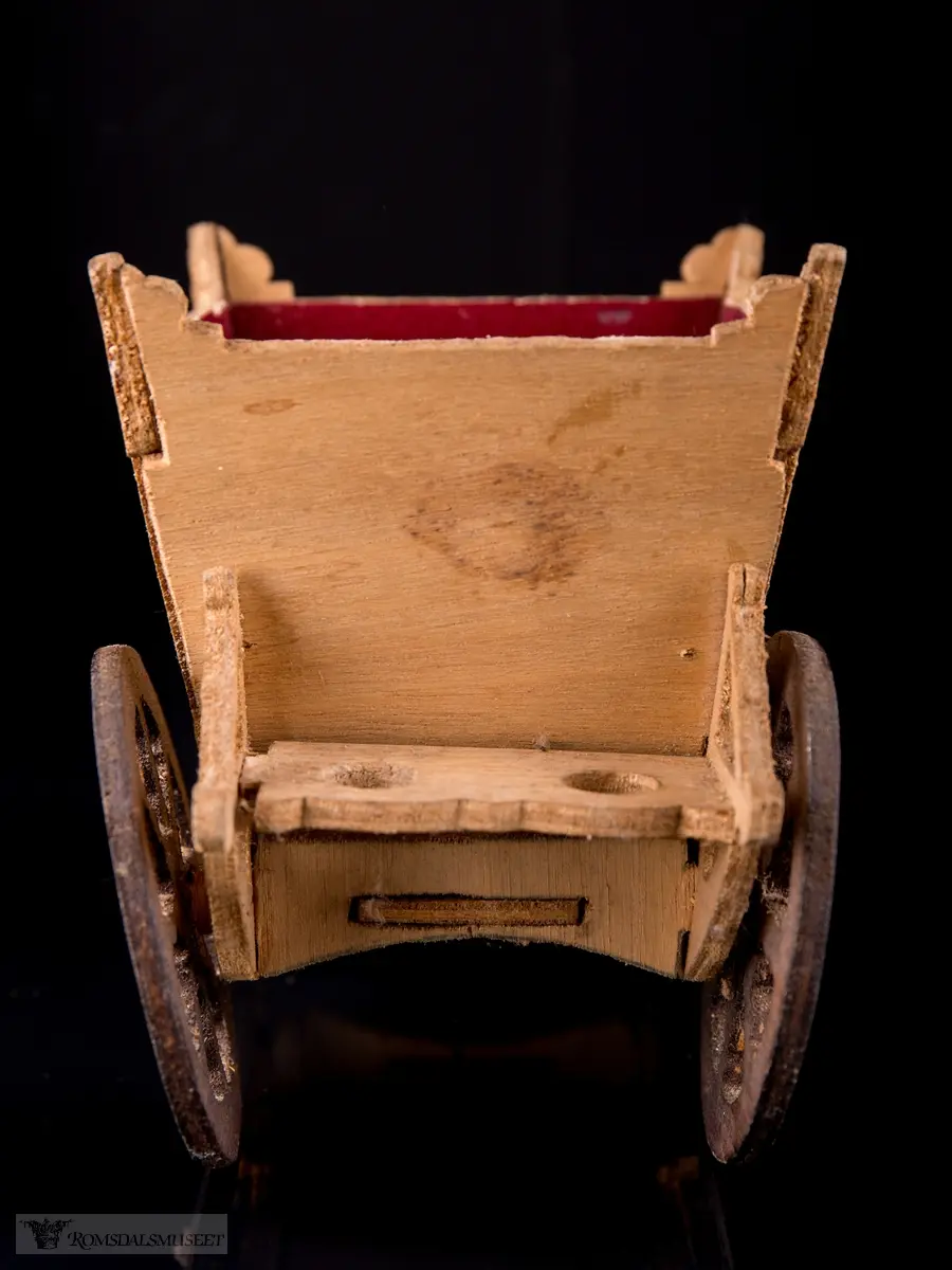 En liten leke- hestevogn i kryssfiner. To store og to små hjul, kledd med rød filt og vokspapir er brukt som vinduer.