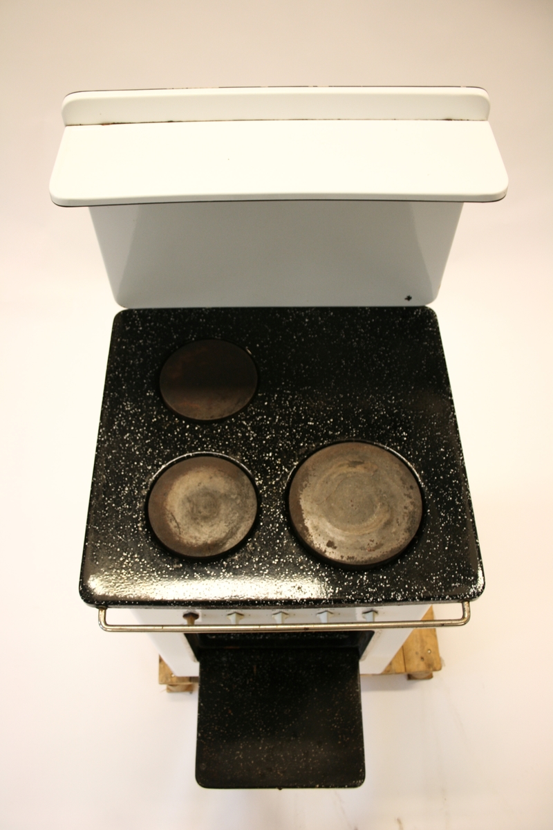 Elektrisk komfyr med bakvegg og tre kokeplater. Stekeovn med termometer på utsiden. I stekeovnen er det to tilhørende stekebrett. Lyspære på bakveggen.