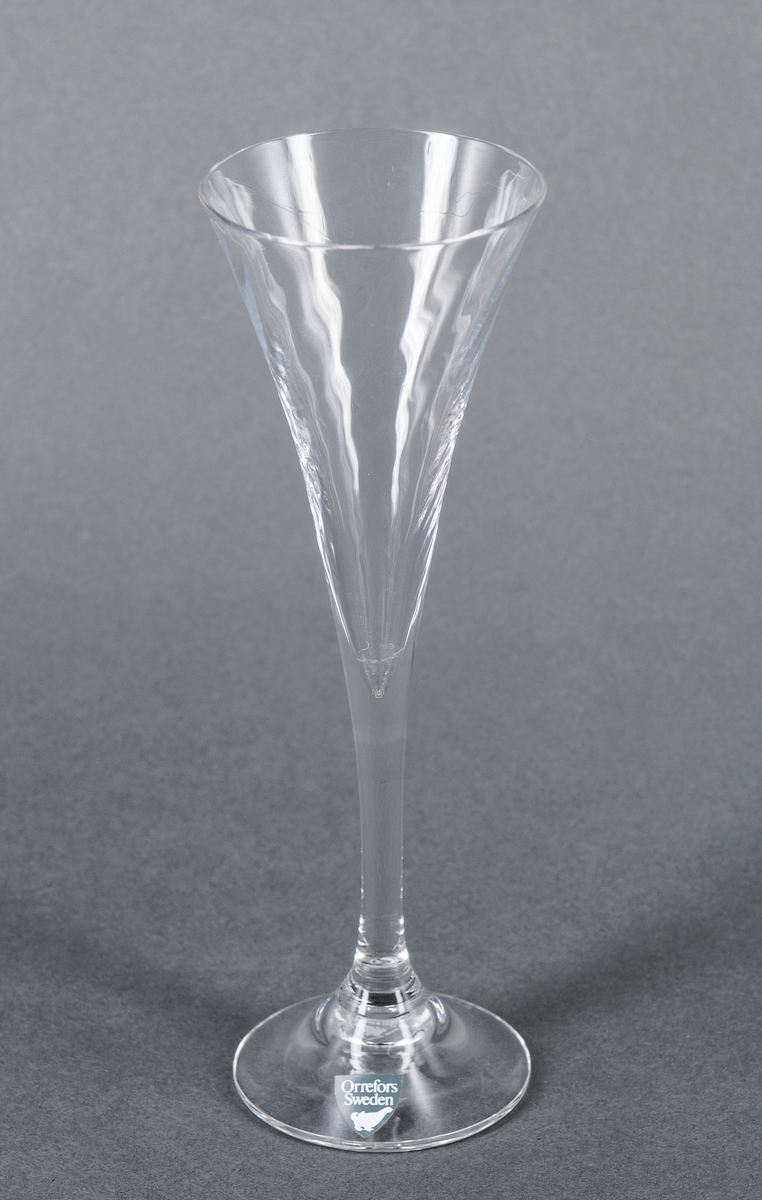 Snapsglas/Sherryglas, ofärgat, modell Helena. Trumpetformad kupa med diagonalställd optik, övergående i högt smalt ben. Grå etikett på foten: Orrefors Sweden