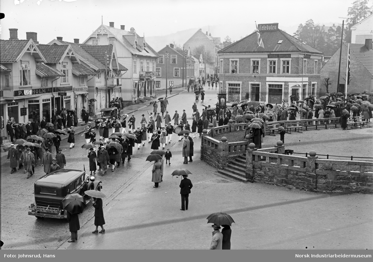 17. mai feiring 1931. Folketog i gaten ved Notodden torg. Tilskuere på fortau. Biler og mennesker gående i gaten. Teledølen lokale i bakgrunnen. Butikkfasader langs Storgata i Notodden sentrum.