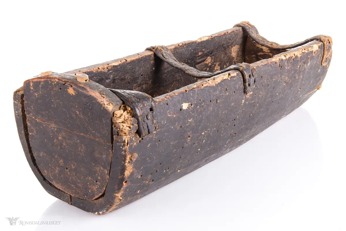 Uthulet trestokk som er høvlet flat i bunnen og har påsatte endestykker som er festet med trenagler. Tre lærremmer er spikret fast på tvers av åpningen. Rester av bek i bunnen.