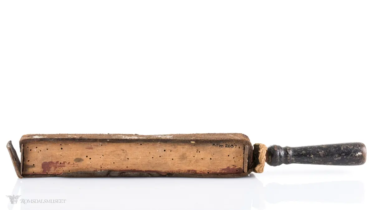 Rektangulær med dreid trehåndtak. Lærrem som går rund hele. Avstivet med metallbeslag foran og bak. Brukt til å stroppe/kvesse barberknivblad før bruk.