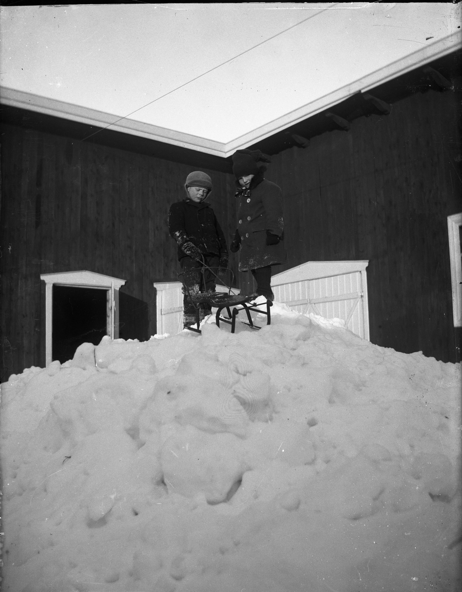 To guttunger og en kjelke på en snøhaug, trolig foran en låve. Hverken guttene eller stedet er identifisert.