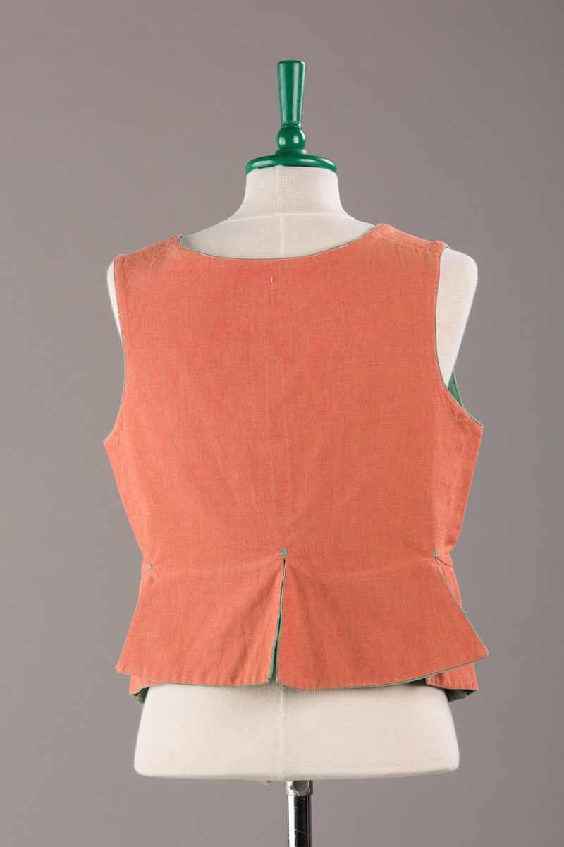 Rustrødt liv med grønn kant og fór, seks skjøter. Håndsydd forsterkning i kontrastfarge øverst i skjøtesplittene. Lukkes med tinnhekte og silkesnor.