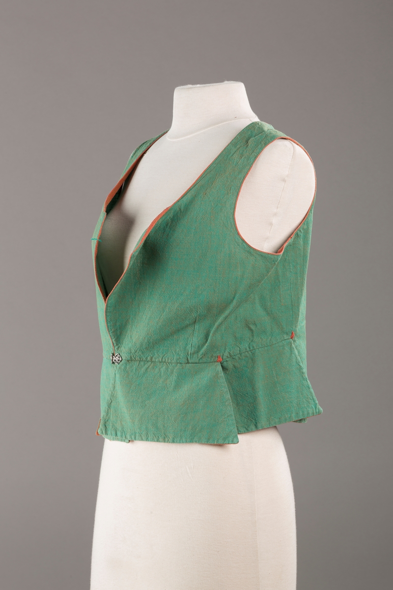 Grønt liv med rustrød kant og fór, seks skjøter. Håndsydd forsterkning i kontrastfarge øverst i skjøtesplittene. Lukkes med tinnhekte.