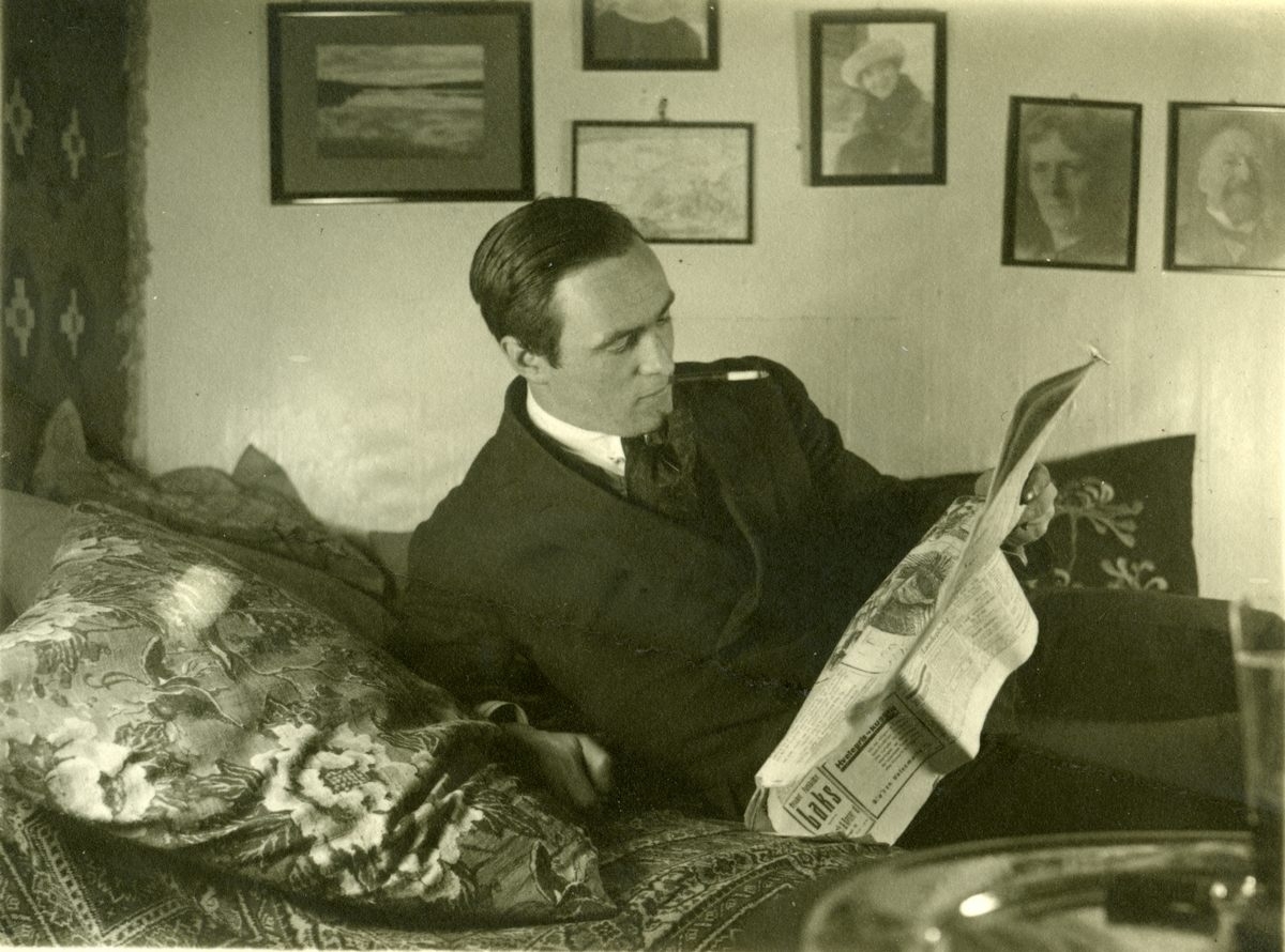 Eyvin Floer Bjørnstad leser avis. I bakgrunnen til h. foto av M.P. Bjørnstad og hans kone.