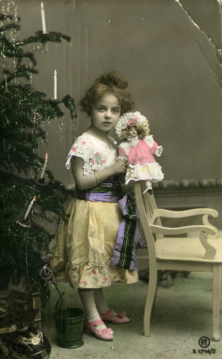 Julekort. Jule- og nyttårshilsen. En jente kledd i en flott kjole står ved siden av et juletre. I armen holder hun en pyntet dukke. Poststempel 24.12.1907.