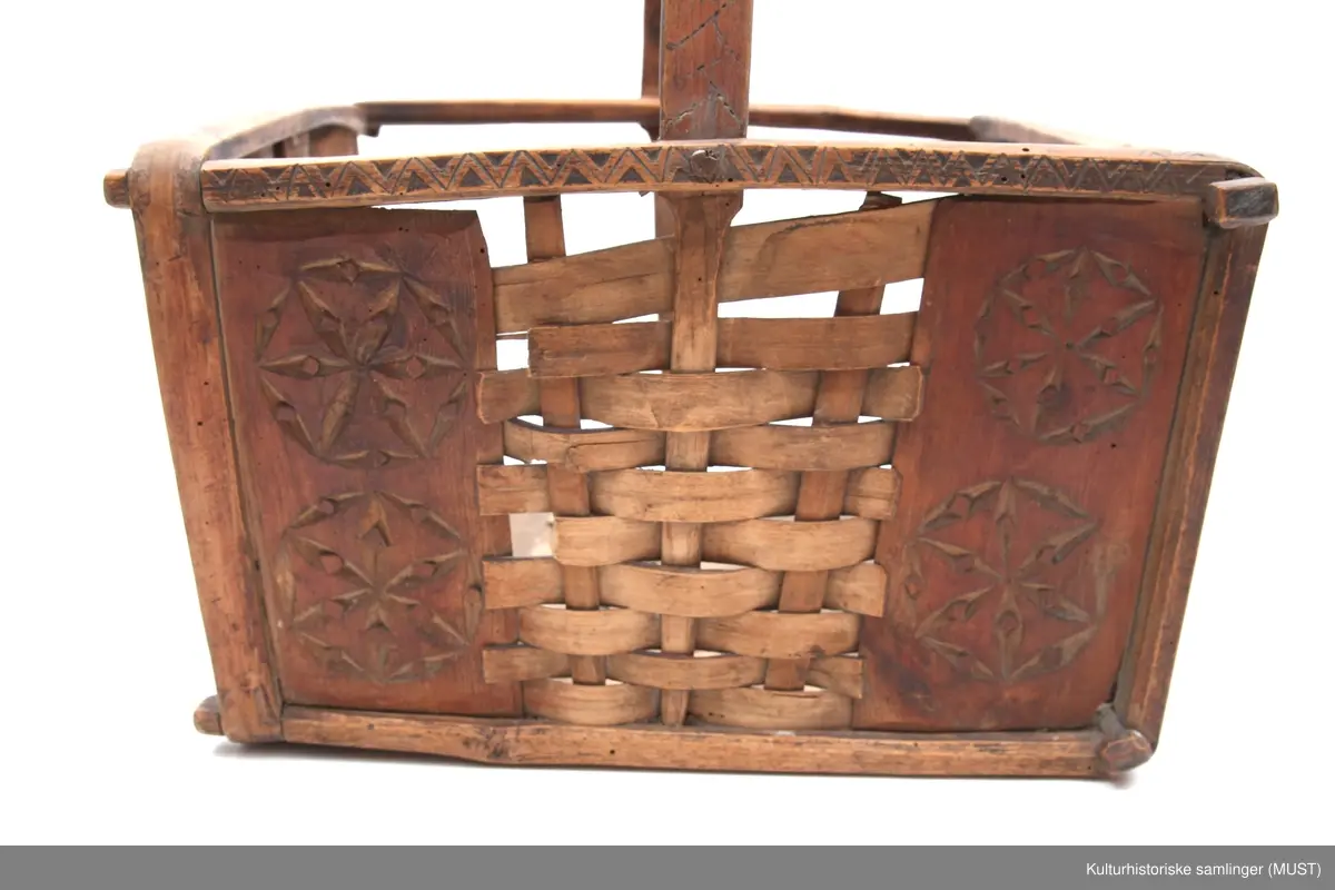 Firkantet trekurv med flettedekor og treskjæring på både kurv og håndtak. Dekoren består av to seksbladete blomster på hver side samt ulike trekantmønstre langs kanten av kurven og håndtaket. En av sidene i kurven mangler.