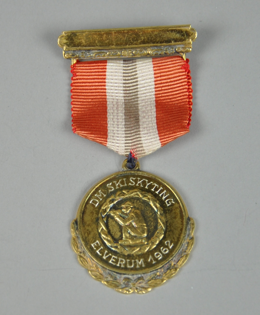 Medalje med laurbærkrans rundt medaljongen og skiskytter som sitter på kne og sikter med geværet. Krans rundt skytteren. Nål med bånd i rødt, hvit og blått (blå farge er falmet).