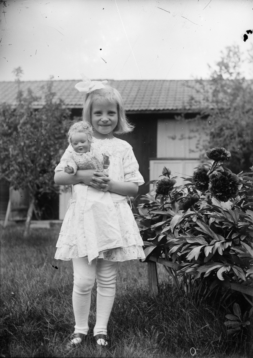 Ruben och Christiane Liljefors dotter Marit står i trädgården vid pionbuske med sin docka i famnen, sannolikt på Svensgården i Dalarna, Sverige
