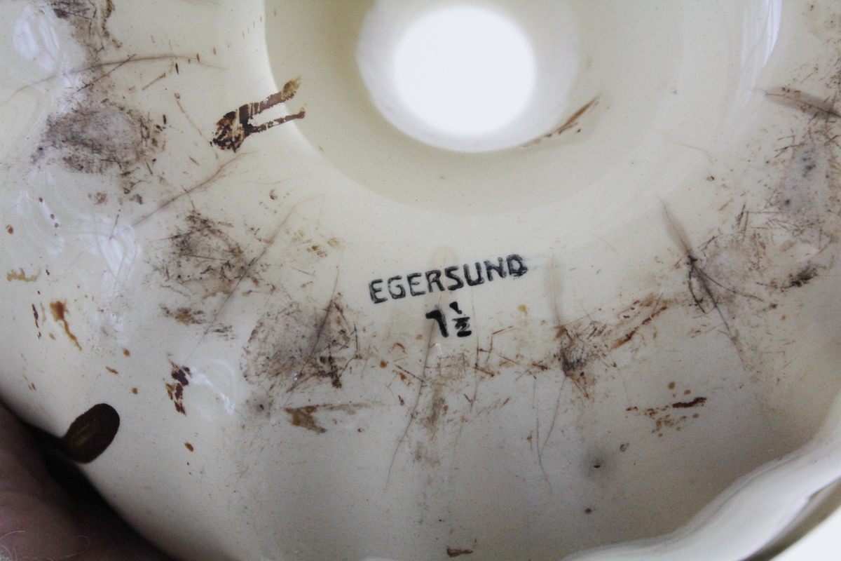 Rund form i ildfast steintøy. Kremgul farge, med brun dekor. Brune stiliserte blomsterkvister på sidene. Rommer halvannen liter.