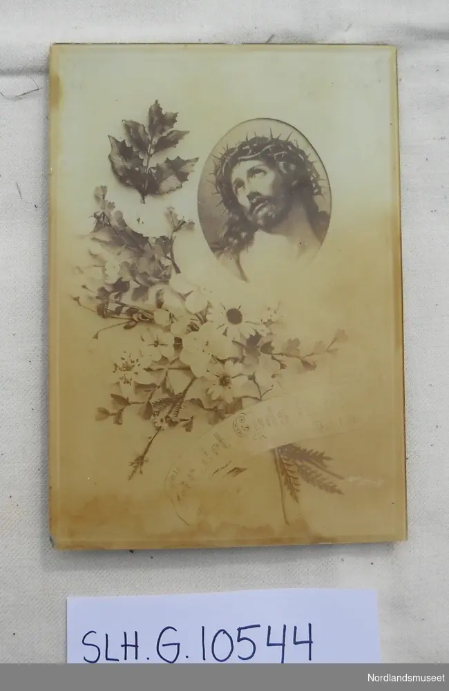 Ramme i glass, påmontert klaff på bakside for at den skal kunne stå. Illustrasjon med bilde av blomsterbukett, ovalt felt med Kristus med tornekrone.